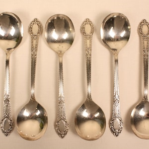 May include: A set of six silver-toned spoons arranged in two alternating rows. Three spoons feature plain, polished oval bowls, while the other three spoons have ornate, embossed floral and scrollwork detailing along the handles and stems.