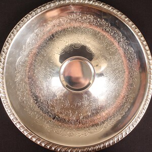 May include: A silver-colored serving tray with an ornate, engraved floral pattern. The tray has a raised, decorative edge and a central, circular handle. The tray is round and reflects light.