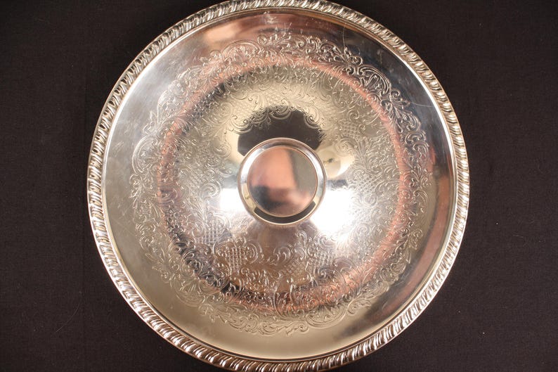 May include: A silver-colored serving tray with an ornate, engraved floral pattern. The tray has a raised, decorative edge and a central, circular handle. The tray is round and reflects light.