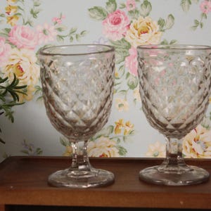 May include: Two clear, vintage-style wine glasses with diamond-patterned detailing. The glasses sit on a dark wooden tray against a floral wallpaper backdrop. The glasses are stemware, perfect for serving wine or other beverages.