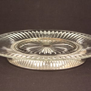 May include: A clear glass cake stand with a silver-colored base. The glass plate has a ribbed design around the edge and a starburst pattern in the center. The base is also ribbed.
