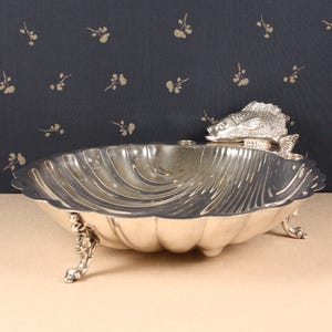 May include: Silver-toned decorative serving dish with a fish-shaped accent. The dish has a scalloped edge and rests on three ornate feet. The fish is positioned on the rim of the dish. The background is a dark blue fabric with a floral pattern.