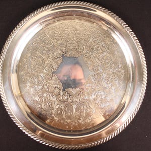 May include: A round, silver-colored serving tray with an ornate, floral pattern in the center. The tray has a rope-like border and is set against a dark background. The reflective surface of the tray shows a distorted reflection.