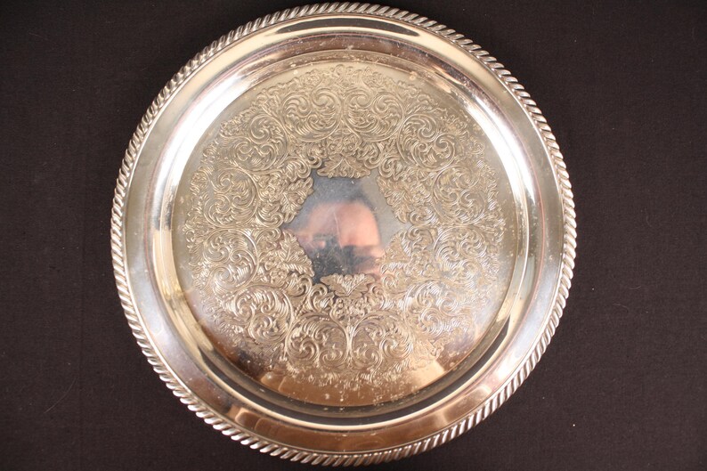 May include: A round, silver-colored serving tray with an ornate, floral pattern in the center. The tray has a rope-like border and is set against a dark background. The reflective surface of the tray shows a distorted reflection.