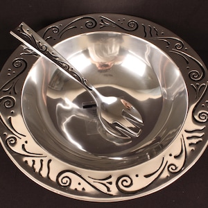 May include: A polished silver-toned metal bowl and a matching serving fork. The bowl features a wide, flat rim decorated with embossed black scrollwork and swirling patterns. The serving fork has a long handle with similar black scrollwork.