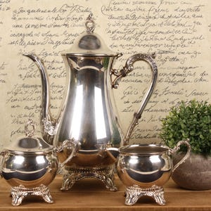 May include: A silver tea set, including a teapot, sugar bowl, and creamer, displayed on a wooden tray. The set has ornate handles and stands on decorative feet. A small potted plant is also on the tray.