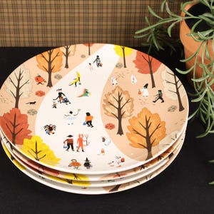 May include: A stack of round ceramic plates featuring a whimsical autumn forest scene with a winding white path. The design includes colorful trees in shades of orange, yellow, and brown, interspersed with small figures dressed in Halloween costumes.