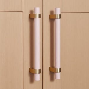 Peut inclure: Gros plan sur une armoire en bois clair à deux portes. Chaque porte est équipée d'une poignée cylindrique rose pâle avec des accents dorés. Les poignées sont fixées avec des ferrures rectangulaires dorées. Des fleurs blanches dans un vase sont visibles au-dessus.