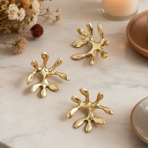 May include: Three gold-colored, abstract-shaped decorative objects are arranged on a marble-patterned surface. The objects have an organic, fluid design. A lit candle, dried flowers, and a cup are also visible.