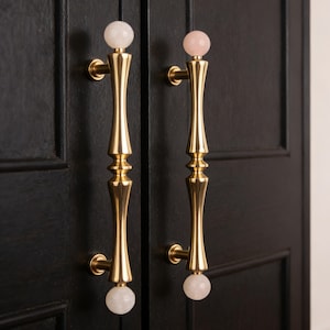 Puede incluir: Dos tiradores dorados para armario con un diseño decorativo. Cada tirador tiene una piedra esférica de color claro en la parte superior e inferior. Los tiradores están montados en una puerta de armario negra.