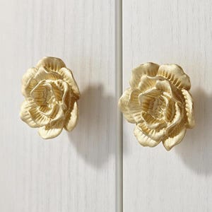 May include: Two gold-coloured, rose-shaped cabinet knobs mounted on a white cabinet door. The knobs have detailed petal designs and are made of a metallic material. The cabinet doors are vertically aligned.