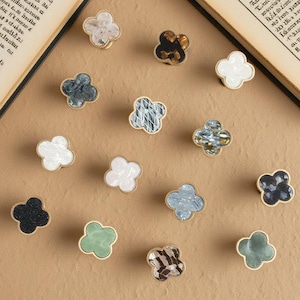 May include: A collection of decorative clover-shaped cabinet knobs in various colors and patterns, including white, black, and green. Each knob has a gold-colored base. The knobs are arranged on a neutral-toned surface.