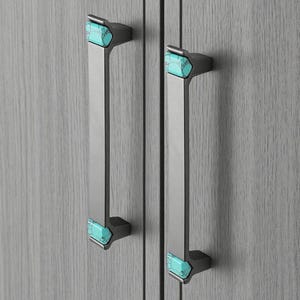 Op de afbeelding: Twee kastgrepen met een rechthoekige, geborstelde metalen handgreep en donkergrijze uiteinden. Elk uiteinde heeft een turquoise, geometrische steen. De grepen zijn gemonteerd op een lichtgrijze houten kast.