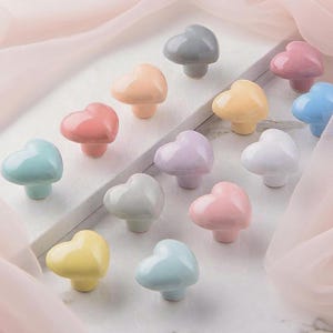 May include: A selection of heart-shaped ceramic cabinet knobs in pastel shades of pink, blue, yellow, and grey. Each knob features a rounded base and a smooth, glossy finish. The knobs are displayed on a white surface.