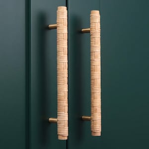 Könnte beinhalten: Zwei Schrankgriffe mit geflochtenem Rattan-Design und messingfarbener Hardware. Die Griffe sind zylindrisch und vertikal ausgerichtet, vor einer dunkel türkisfarbenen Schranktür. Das Rattan hat eine natürliche, hellbraune Farbe.