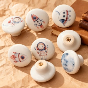 May include: A collection of white ceramic cabinet knobs with space-themed illustrations. Designs include rockets, astronauts, and a moon. Each knob has a brass-colored screw fitting on the back. These knobs are perfect for adding a whimsical touch to furniture.