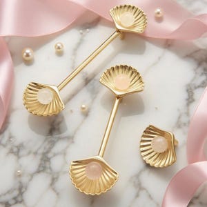 May include: Gold-colored seashell-shaped cabinet pulls and knobs with a light pink gemstone center. The hardware is arranged on a marble surface with scattered pearls and pink ribbon, creating an elegant aesthetic.
