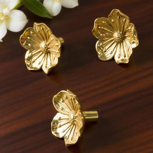 May include: Three gold-colored flower-shaped cabinet knobs. Each knob features a detailed floral design with petals and a central stamen. The knobs are set against a dark wood surface, with white flowers in the background.