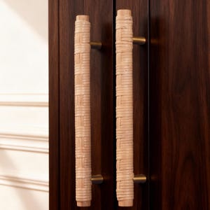 Może przedstawiać: Dwa uchwyty do szafek z uchwytami z plecionego jasnobrązowego rattanu i mosiężnym osprzętem. Uchwyty są zamontowane na ciemnobrązowych drewnianych drzwiach szafki. Drzwi szafki są zamknięte.