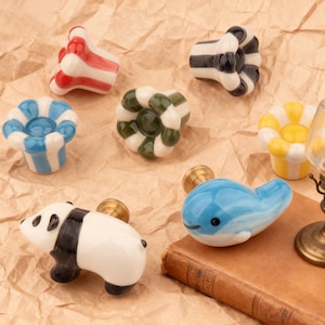 Pode incluir: Uma coleção de puxadores de cerâmica decorativos com vários designs. Os puxadores apresentam um panda, uma baleia azul e padrões listrados em vermelho, preto, azul, verde e amarelo. Cada puxador tem uma base de cor latão.