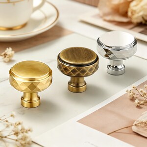May include: Three decorative cabinet knobs in gold, bronze, and silver. Each knob has a round top with a diamond pattern around the base. The gold knob has a bright finish, the bronze knob has a matte finish, and the silver knob has a shiny finish.