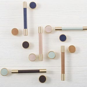 Könnte beinhalten: Eine Sammlung von Schrankbeschlägen, darunter zylindrische Griffe und runde Knöpfe. Die Griffe haben goldfarbene Metallakzente und sind mit Leder in verschiedenen Farben wie Blau, Rosa und Braun umwickelt. Die Knöpfe sind farblich passend.