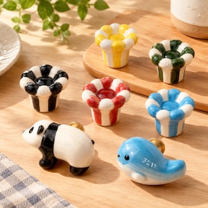 May include: A collection of ceramic cabinet knobs including five flower-shaped knobs in yellow, green, black, red, and blue stripes, one panda-shaped knob, and one blue whale-shaped knob. The panda and whale knobs feature the engraved text 3219.