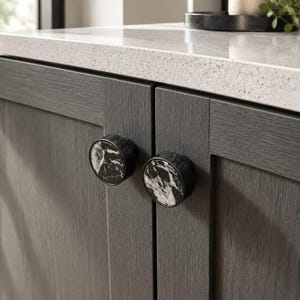 May include: Two circular cabinet knobs featuring a black and white marble inlay set within a matte black metal frame. Each knob displays the engraved number 3219 on the side of the metal rim, mounted on dark gray wood-grain cabinet doors.