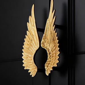 May include: Gold-colored, ornate angel wing door handles mounted on a black door. The wings are detailed with individual feathers and are symmetrical. The hardware adds a decorative touch to the door.