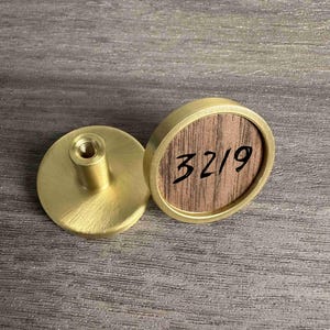 May include: Two circular cabinet knobs featuring a brushed gold metal finish. One knob shows a flat, threaded mounting post, while the other displays a circular wood grain inlay with the black handwritten text 3219 centered on the surface.