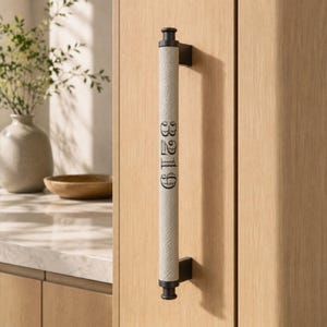 May include: A vertical cabinet handle featuring a textured, light beige cylindrical grip with the black text 6129 printed vertically. The handle is secured by two dark brown metal brackets attached to a light-colored wooden cabinet door surface.