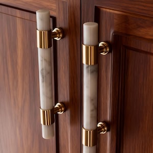 May include: Cabinet door handles with a cylindrical design. The handles have a white marble centre with gold-coloured metal accents. They are attached to a dark brown wooden cabinet door.