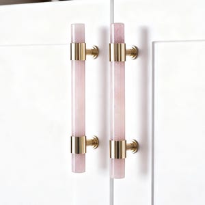 以下が含まれることがあります： 淡いピンク色の長方形のセンターと金色の金属アクセントが付いた2つのキャビネットプル。プルは白いキャビネットドアに取り付けられています。プルは円筒形で、モダンなデザインです。