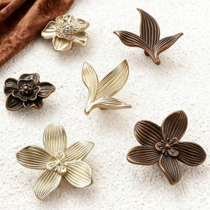 Op de afbeelding: Een assortiment decoratieve kastknoppen in verschillende bloemen- en bladvormen. De knoppen zijn in goud- en bronskleuren, met gedetailleerde texturen en vormen. De opstelling omvat hardware in bloem- en bladvorm.