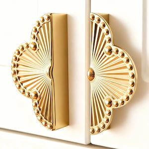 Op de afbeelding: Twee gouden kastgrepen met een waaiervormig ontwerp. Elke greep heeft een reeks kleine, ronde accenten langs de rand en een centraal, afgerond detail. De grepen zijn gemonteerd op een witte kastdeur.