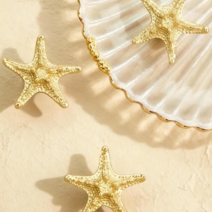 Puede incluir: Tres decoraciones de estrellas de mar doradas están dispuestas cerca de un plato decorativo de concha con un borde dorado. Las estrellas de mar tienen texturas detalladas y están sobre un fondo neutro, evocando un tema costero.
