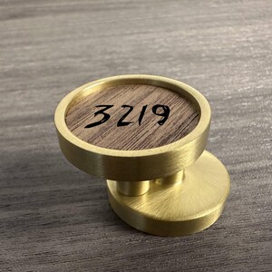 May include: A circular brass knob featuring a central inlay of brown wood grain. The wood surface is marked with the black handwritten number 3219. The knob has a brushed metallic finish and is mounted on a matching circular brass base plate.