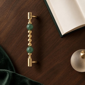 May include: A gold-toned metal cabinet handle featuring a vertical design with two large green spherical beads and three smaller gold spherical beads in the centre. The top of the handle is engraved with the black text 3219.