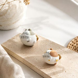 May include: Two rounded cabinet knobs shaped like stylised cats, one with grey and white markings and the other with brown and white markings. Each knob features the number 3219 printed in black on the front and a gold-toned metal mounting base.