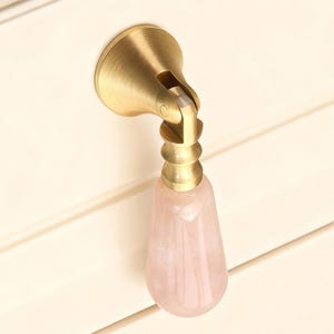 May include: A cabinet pull with a brass finish and a teardrop-shaped rose quartz handle. The pull is mounted on a cream-colored cabinet door. The brass hardware has a decorative, articulated design.