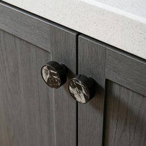 Può includere: Primo piano di due pomelli per armadi. I pomelli sono neri con un design simile al marmo nelle tonalità del bianco e del marrone. I pomelli sono attaccati a un'anta grigia. Un piano di lavoro bianco è visibile sullo sfondo.