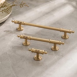 May include: Three brass-toned metal cabinet pulls of varying lengths, each featuring a cylindrical bar with rounded finials and a central engraved marking of the number 3219. The hardware includes circular mounting bases for attachment to furniture.