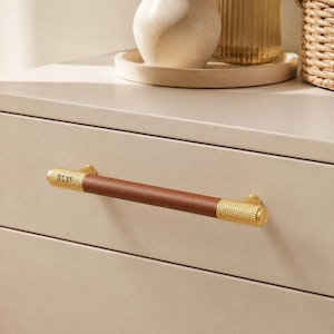 May include: A drawer pull featuring a central brown leather grip flanked by two textured gold-toned metal ends. The hardware is mounted horizontally on a smooth, light beige drawer front, showcasing a combination of smooth leather and knurled metal.
