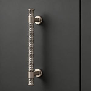 Pode incluir: Um puxador de armário em níquel escovado com um padrão tecido na pega. O puxador é montado numa porta de armário cinza escuro. O hardware tem uma estética moderna e industrial.