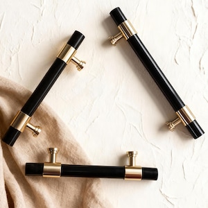 May include: Four black cabinet pulls with gold-coloured accents. The pulls have a cylindrical shape with a gold band in the middle and gold-coloured mounting hardware. The pulls are arranged on a white textured surface with a beige cloth.