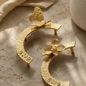 May include: Two gold-colored metal drawer pulls with a curved, hammered texture. One pull features a butterfly ornament, while the other features a flower ornament. Both pulls have the number 3219 engraved in black on the curved surface.