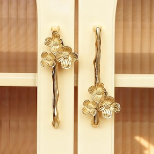 Puede incluir: Un par de tiradores dorados para armario con diseño floral. Cada tirador presenta tres flores y un tallo curvo. Están montados en una puerta de armario color crema.
