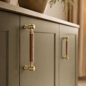 May include: Close-up of a cabinet with olive green doors and brass hardware. The cabinet features two vertical brass handles with a dark wood center. The handles are mounted on the cabinet doors, providing a stylish and functional design element.