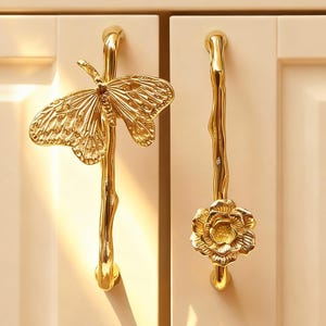 May include: Gold-coloured cabinet pulls with nature-inspired designs. One pull features a butterfly, the other a flower. Both are mounted on a cream-coloured cabinet door. The pulls have a detailed, textured finish.