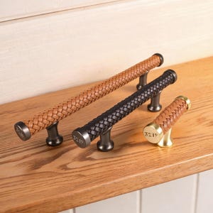 May include: Three cabinet pulls featuring braided leather textures. Two long pulls have dark metal ends engraved with 3219, one in tan leather and one in black. A smaller, single-post knob features tan braided leather and a gold-toned metal end.